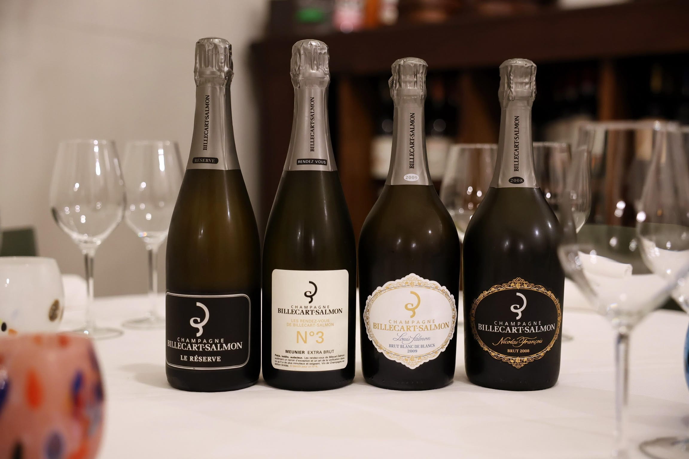 Quattro bottiglie di Champagne Billecart-Salmon allineate su un tavolo, con diversi bicchieri da vino sullo sfondo. Le etichette visibili includono Le Réserve, N°3 Meunier Extra Brut, Louis Salmon Brut Blanc de Blancs 2009 e Nicolas François Brut 2008